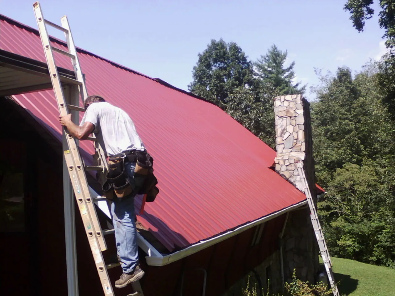 Roofer on a ladder beside a red metal roof and stone chimney — Soffit & Fascia Repair by Roofing Brothers in Cypress Inn